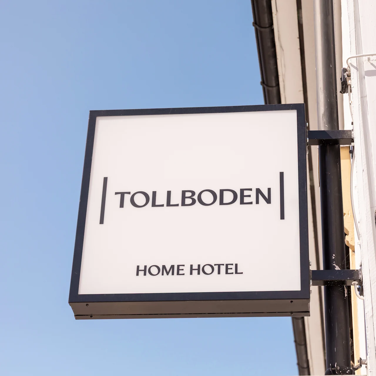 Et kvadratisk skilt viser "TOLLBODEN" og "HOME HOTEL" i tykke bokstaver, montert på en hvid bygning mod en klart blå himmel.