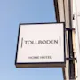 Et kvadratisk skilt viser "TOLLBODEN" og "HOME HOTEL" i tykke bokstaver, montert på en hvid bygning mod en klart blå himmel.