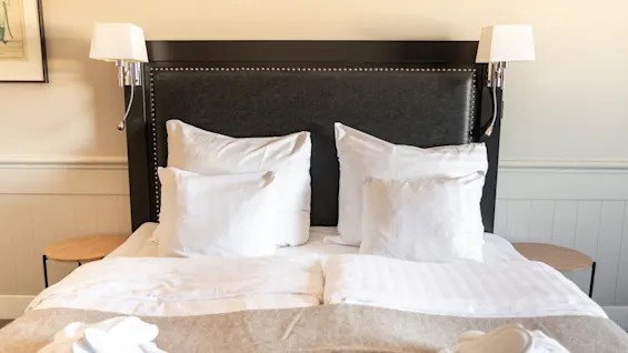 En bekväm och inbjudande hotellsäng med krispiga vita lakan, en mörk sänggavel och sänglampor, vilket antyder en avkopplande vistelse.