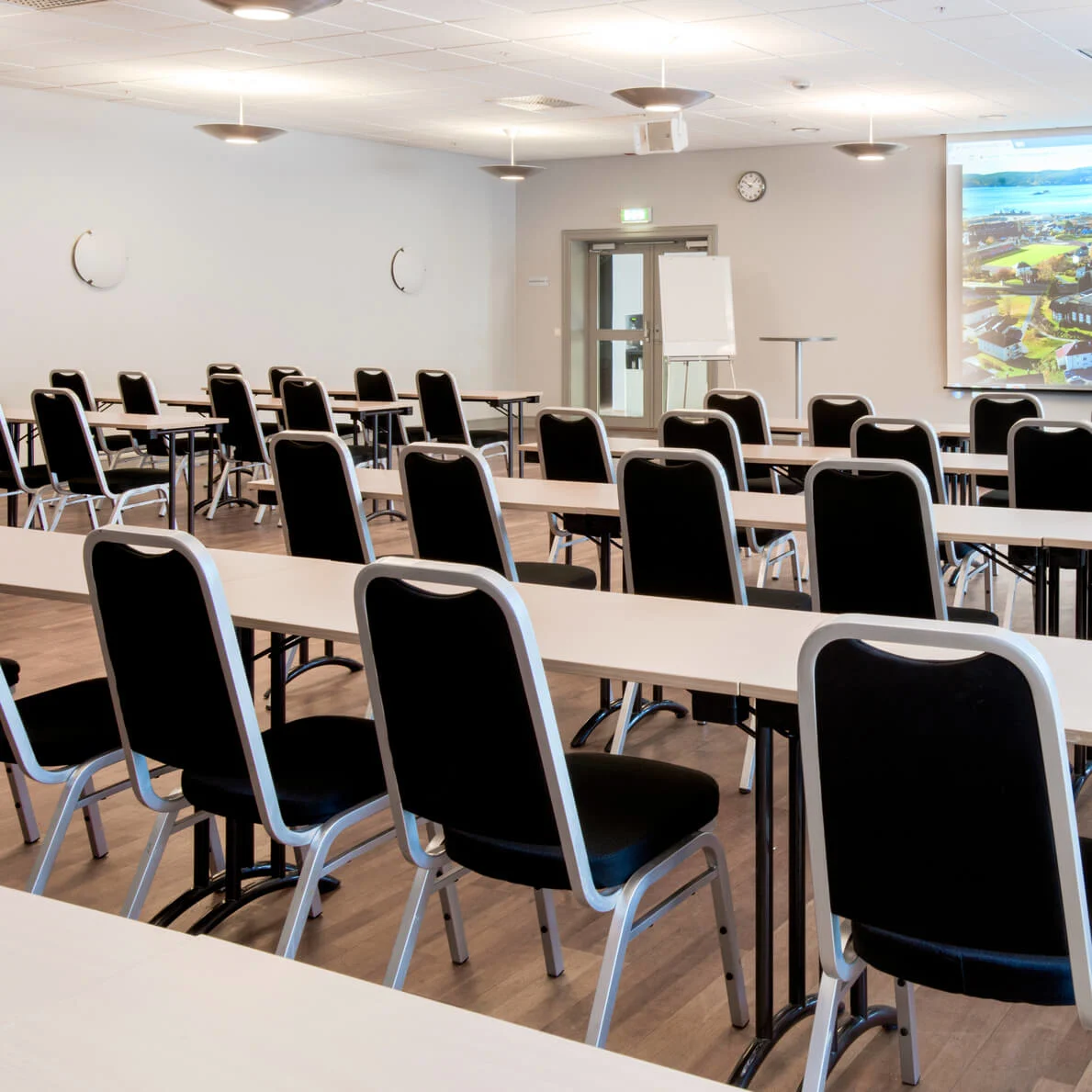 Rader av svarta stolar vända mot en projektorduk i ett tomt konferensrum.