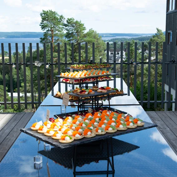 Etasjedelt utendørs matbuffé fylt med gourmet-fingermat på en terrasse med fantastisk utsikt over fjorden.