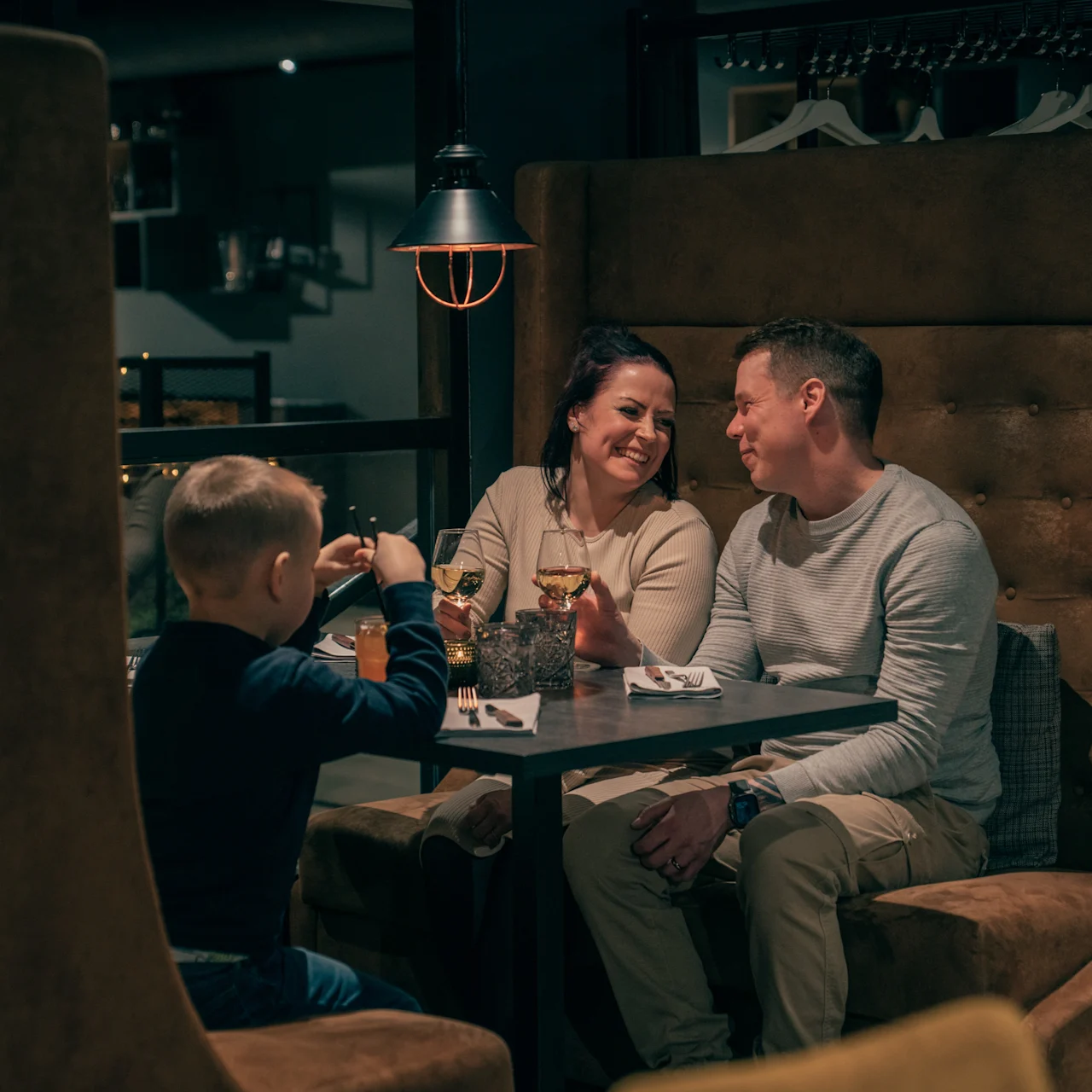 A family sits at a restaurant booth, smiling and enjoying drinks under warm lighting. Coats hang on racks in the softly lit background.