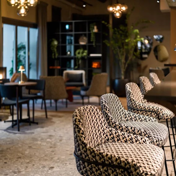 Hyggelig hotellbar med mønstrede barstoler langs en mørk disk, varm belysning og loungemøbler i bakgrunnen.