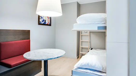 Et moderne rum med køjeseng, rød polstret bænk, marmorbord, abstrakt indrammet maleri og stor loftslampe. Neutrale vægge og trægulv skaber en minimalistisk indretning.