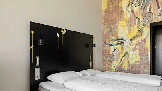 Bed with white linens rests against a black headboard adorned with artistic yellow decorations. A vibrant mural with a dynamic figure and flowers decorates the adjacent wall.