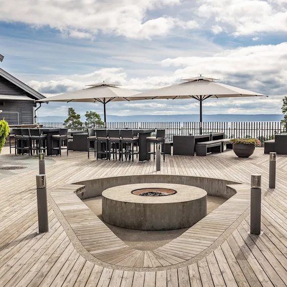 Oversiktsbilde av en stor utendørs terrasse med sittegrupper, bålpanne og utsikt over vannet.