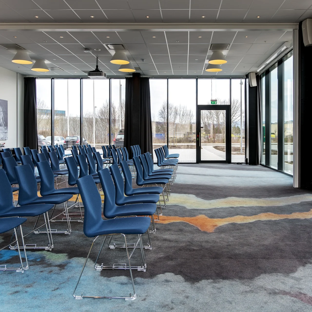 Rows of blue chairs face forward in a spacious, modern conference room. The room features large glass windows showing trees outside, and colorful carpet patterns on the floor.