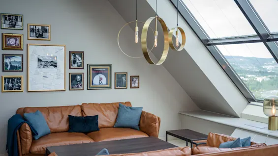 Brune skinnsofaer står i et koselig loft med blå puter, under et skråvindu. Moderne pendellamper henger over, og innrammede bilder pryder den tilstøtende veggen.