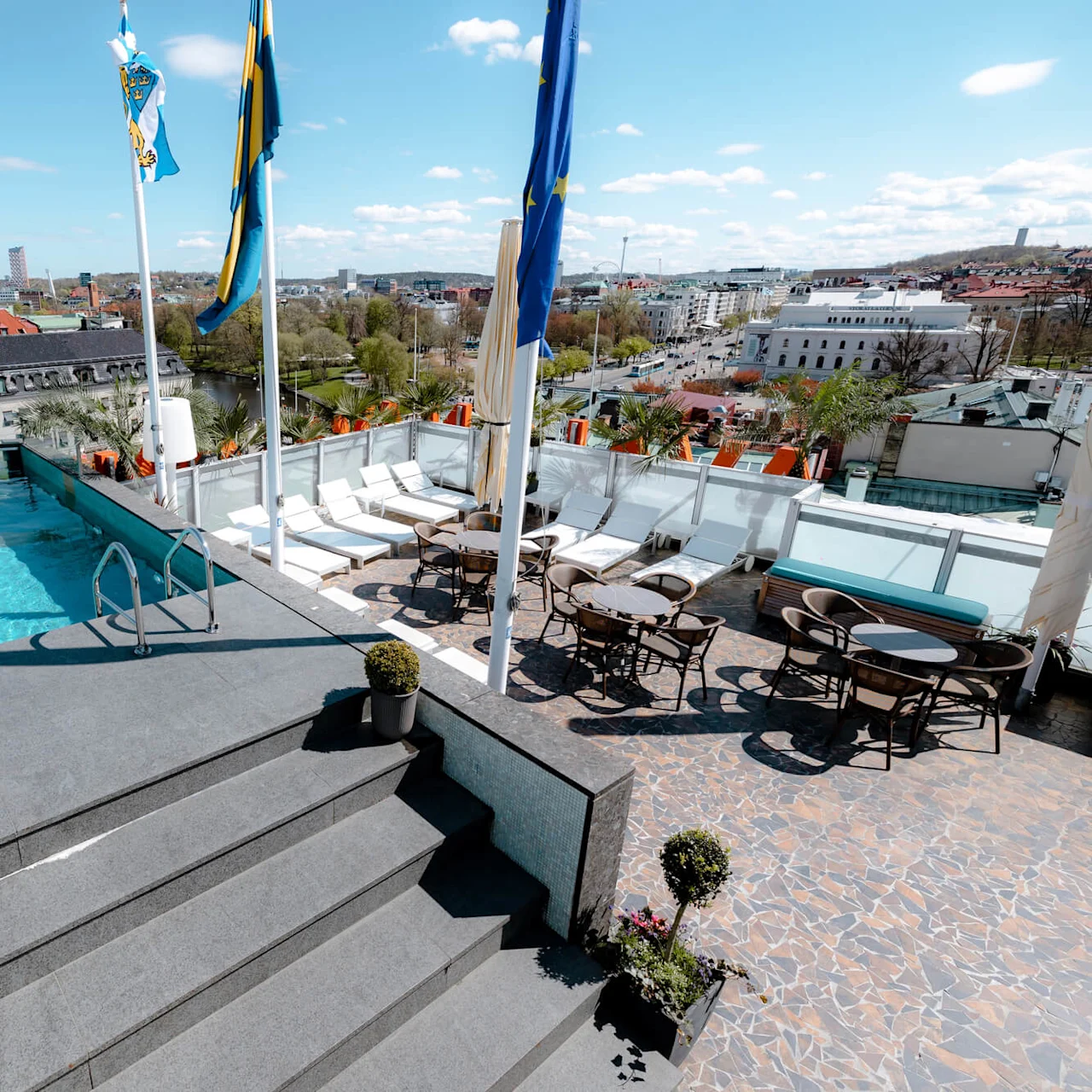 A rooftop infinity pool overlooks a cityscape under a blue sky. Flags, lounge chairs, and tables with chairs surround the pool, providing a relaxing urban environment.