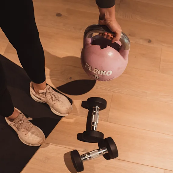 Person på en treningsmatte som holder en rosa kettlebell, med to hantler på tregulvet.