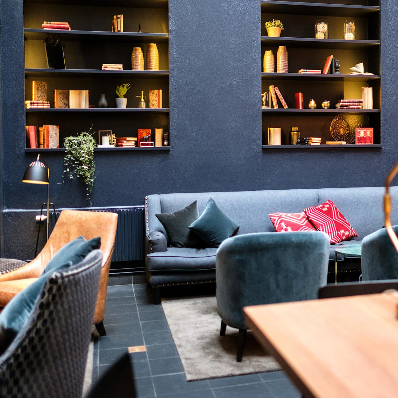 A cozy lounge area with plush sofas, pillows, a bookshelf, and a prominent green desk lamp illuminating a wooden table.