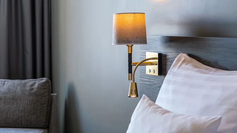 Wall-mounted lamp illuminating, attached above a bed's headboard with white pillows in a softly lit room featuring gray walls and dark curtains.