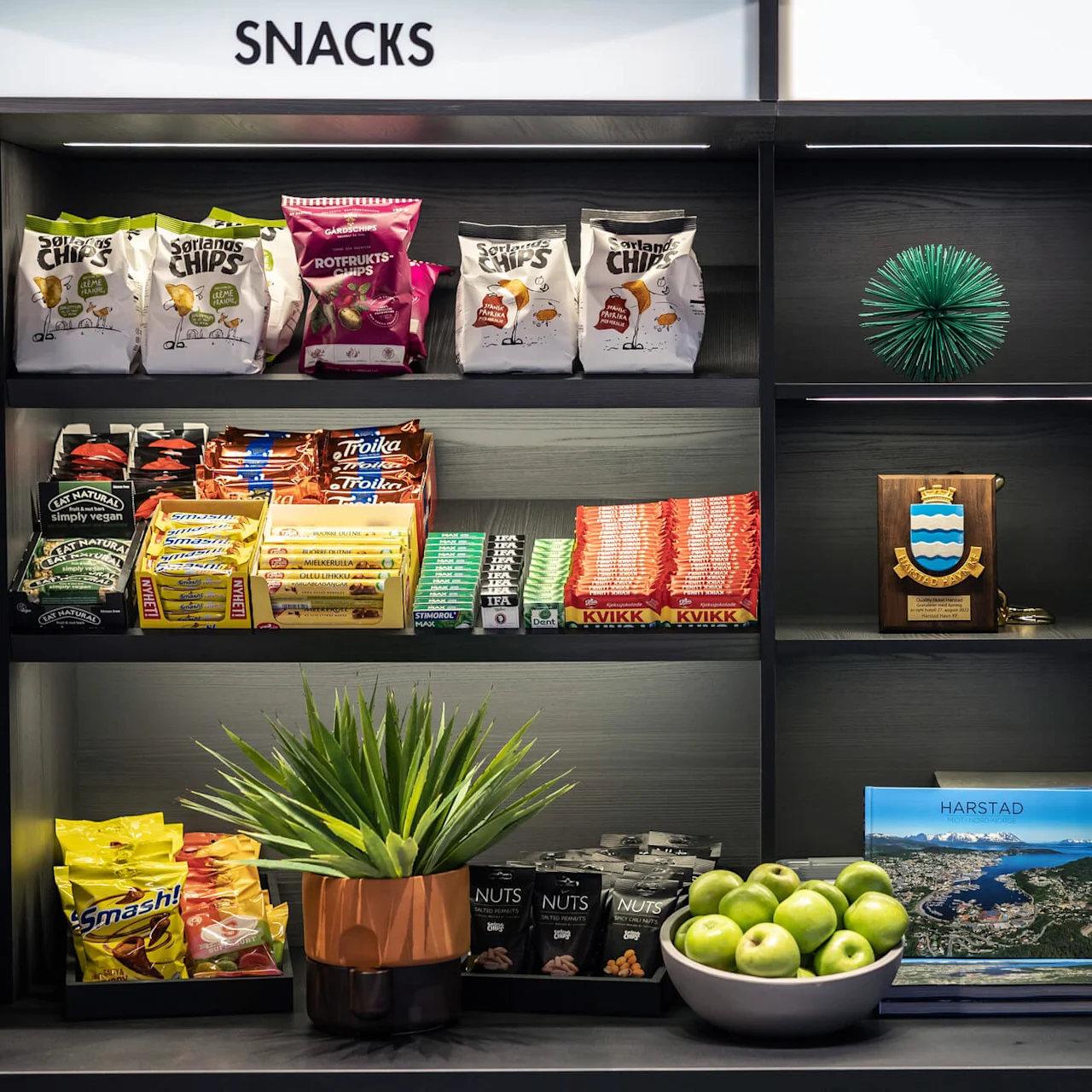 Snacks og kaffestation med forskellige chips, chokolade og nødder på hylder. En kaffemaskine, grønne æbler i en skål og en lille plante pynter området. Tekst: "SNACKS," "COFFEE," "HARSTAD."