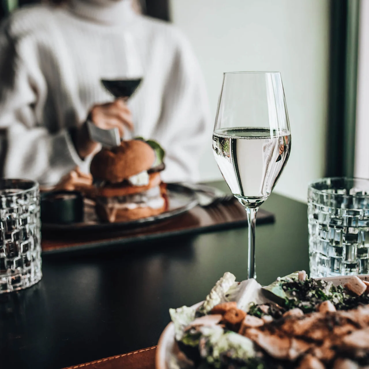 Vinglas står på et restaurantbord. En person i hvid sweater holder en drink nær en burger, omgivet af salat og glas.