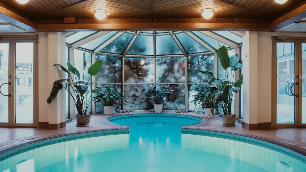 Ett inbjudande inomhuspoolområde med bubbelpool, omgivet av frodiga gröna växter och stora fönster, under ett varmt trätak.