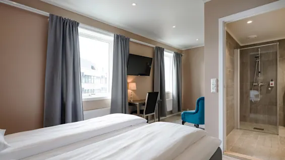 A neatly made double bed sits in a hotel room with gray curtains and a TV. Adjacent is an open bathroom with a glass shower. The room has beige walls.