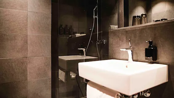 Et moderne bad med vask, spejl og en vægmonteret bruser i en neutral, flisebelagt omgivelse.