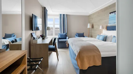Et pent organisert hotellrom med en seng med blått og hvitt sengetøy, et skrivebord i tre med en stol og en TV på veggen. To blå stoler står ved et vindu med blå gardiner.