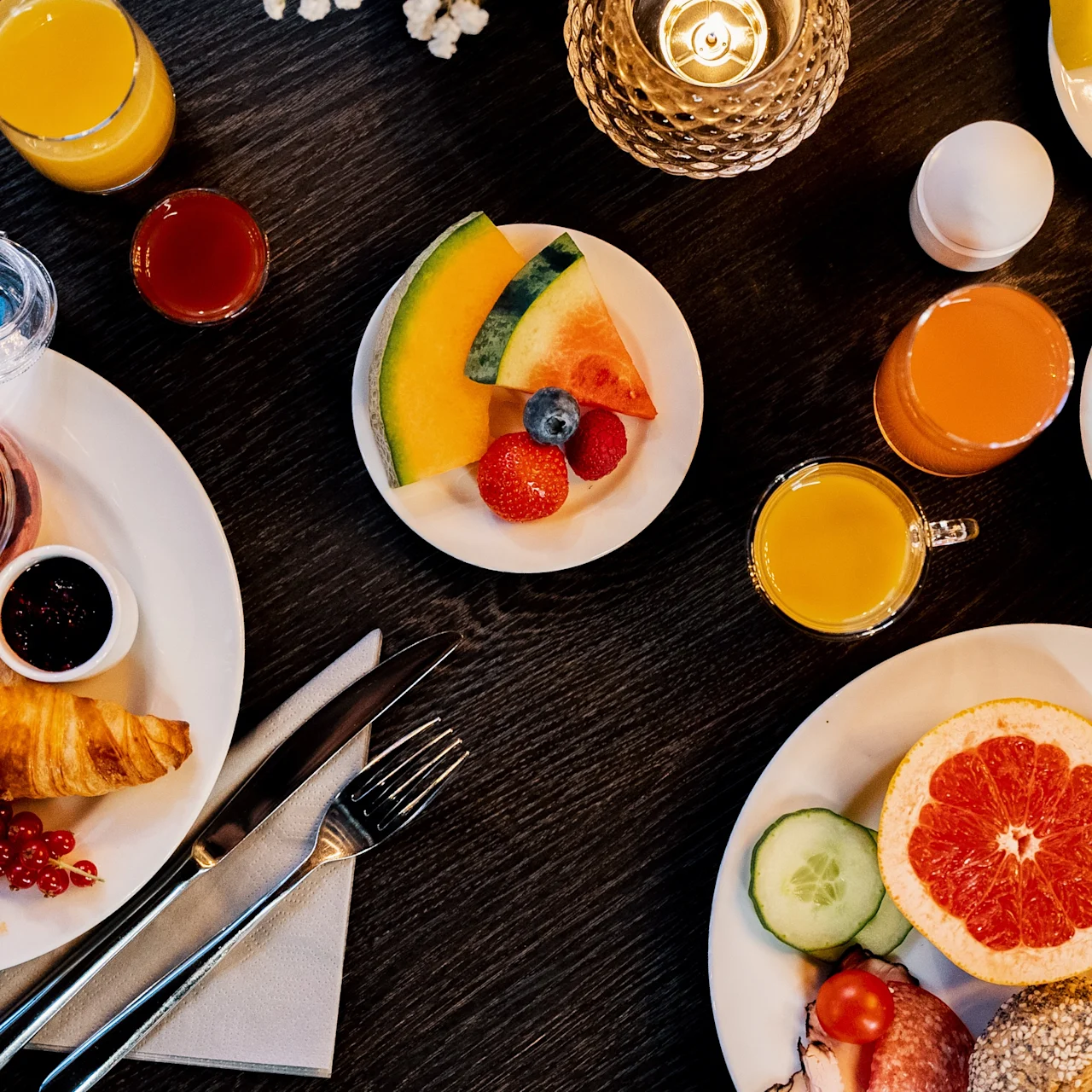 En mängd frukostmat, inklusive croissanter, bär, skivad grapefrukt och juicer, är arrangerade på vita tallrikar på ett mörkt träbord med bestick och ett ljus.