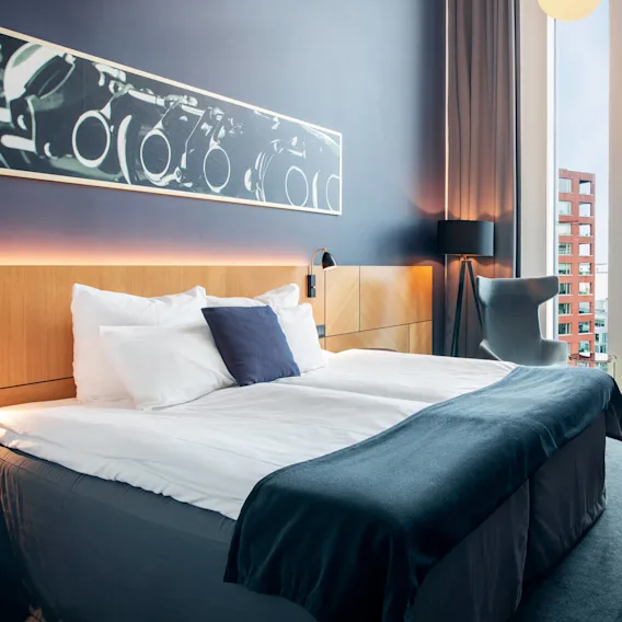 En säng med vita sängkläder är placerad i ett modernt hotellrum med ett stort fönster; dekorativ konst och möbler ger en stilfull atmosfär.