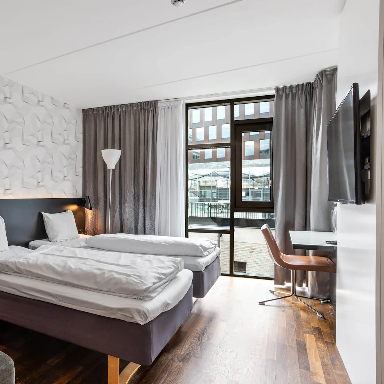 A modern bedroom with two separate beds, textured wallpaper, floor-to-ceiling window, and wooden floor in an urban setting.