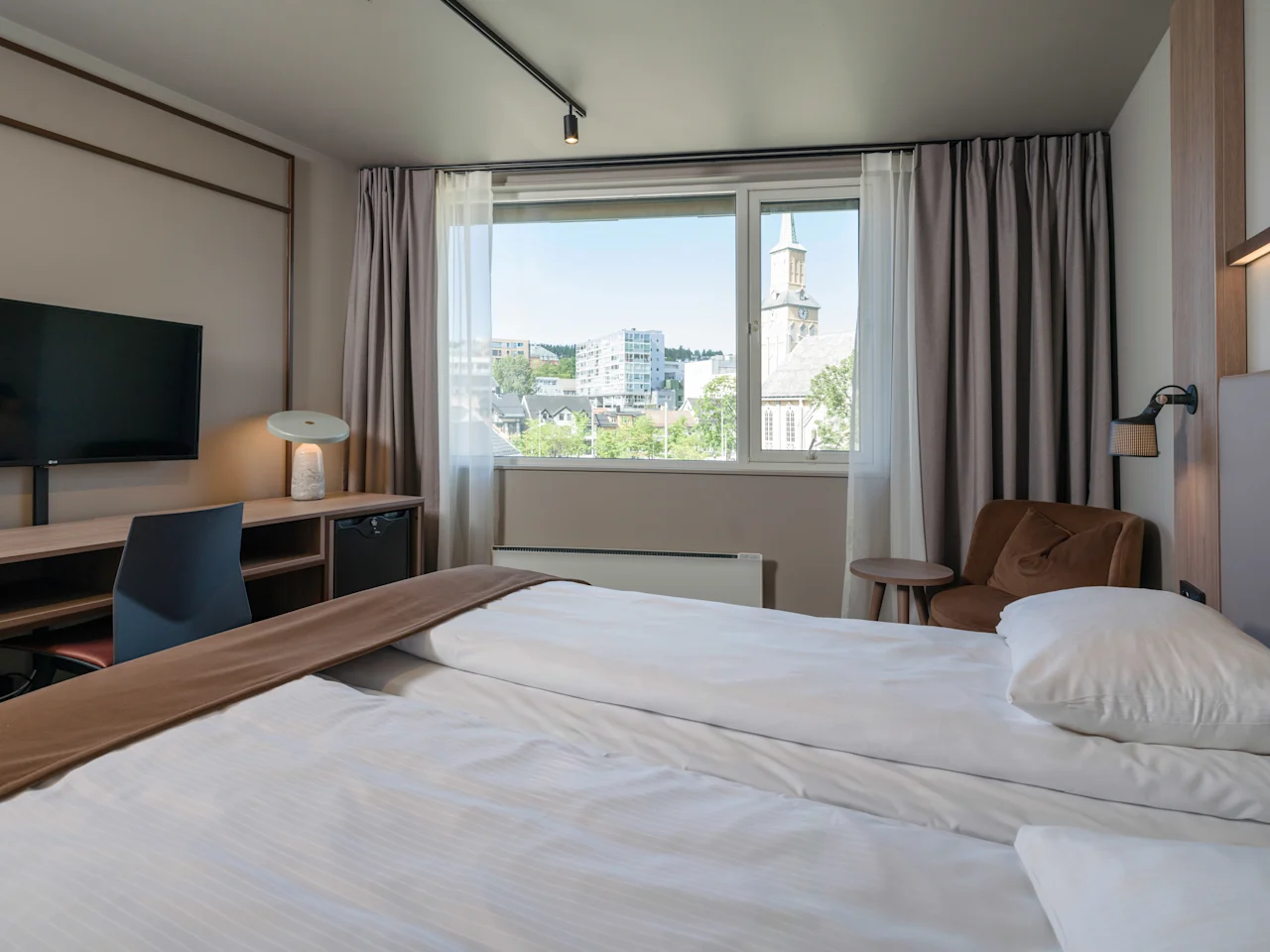 En seng er pænt redt med hvidt sengetøj på et hotelværelse. Et skrivebord med lampe og TV står i nærheden. Et vindue viser udsigt over en by med et kirketårn.