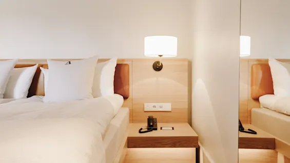 Mysigt hotellrum med en bäddad säng, sänggavel i trä, nattduksbord och en vägglampa som speglas i en spegel.