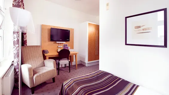 A well-lit hotel room, featuring a striped armchair, small desk with a TV, and a framed artwork on the wall.