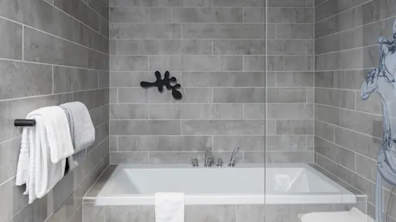 A rectangular bathtub is surrounded by gray tiled walls, with a white towel draped over its edge. Towels hang on a nearby rack, enclosed by glass.
