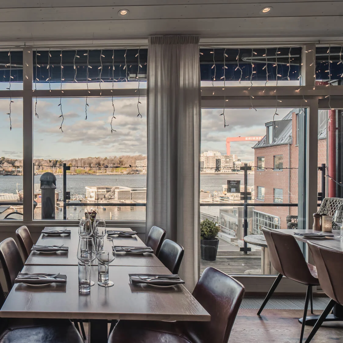 Dining tables set with glassware and napkins face large windows overlooking a waterfront. Twinkling lights decorate the window frames. An industrial bridge and buildings are visible in the distance.