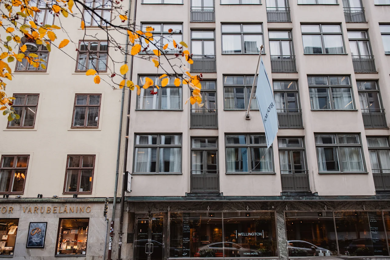 Eksteriør af Home Hotel Wellington i Stockholm med flag og efterårsblade. Stueetagen har store vinduer.