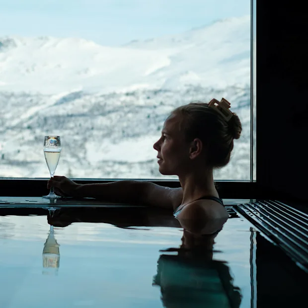 En person slapper av i et basseng, holder et champagneglass, med snødekte fjell synlige gjennom et stort vindu.