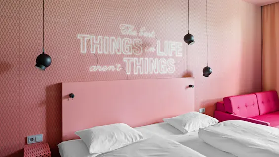 Et rosa soverom med hvitt sengetøy, svarte hengelamper og en rosa sofa. Teksten på veggen lyder: "The best THINGS in LIFE aren’t THINGS."