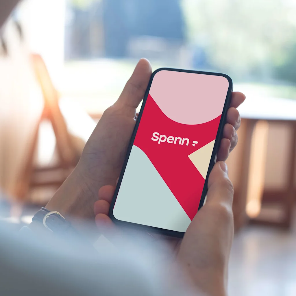 Person holder en smarttelefon med synlig skjerm som viser en grafikk med ordet "Spenn" i en lys og moderne design, innendørs med uskarp bakgrunn.
