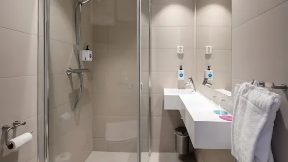 Et moderne bad har en glasslukket dusj, veggmontert dusjhode, hvit vask med to dispensere, en håndklestang med et håndkle og overlys. Veggene er flislagt i beige, og det er en søppelbøtte under vasken.