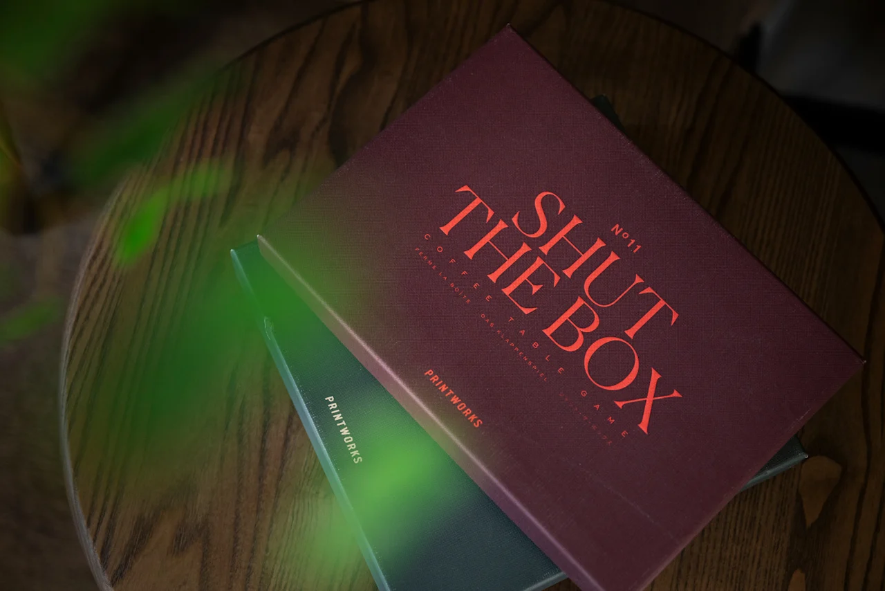 En rödbrun låda märkt SHUT THE BOX N11 COFFEE TABLE GAME FERME LA BOITE DAS KLAPPENSPIEL vilar på en träyta.