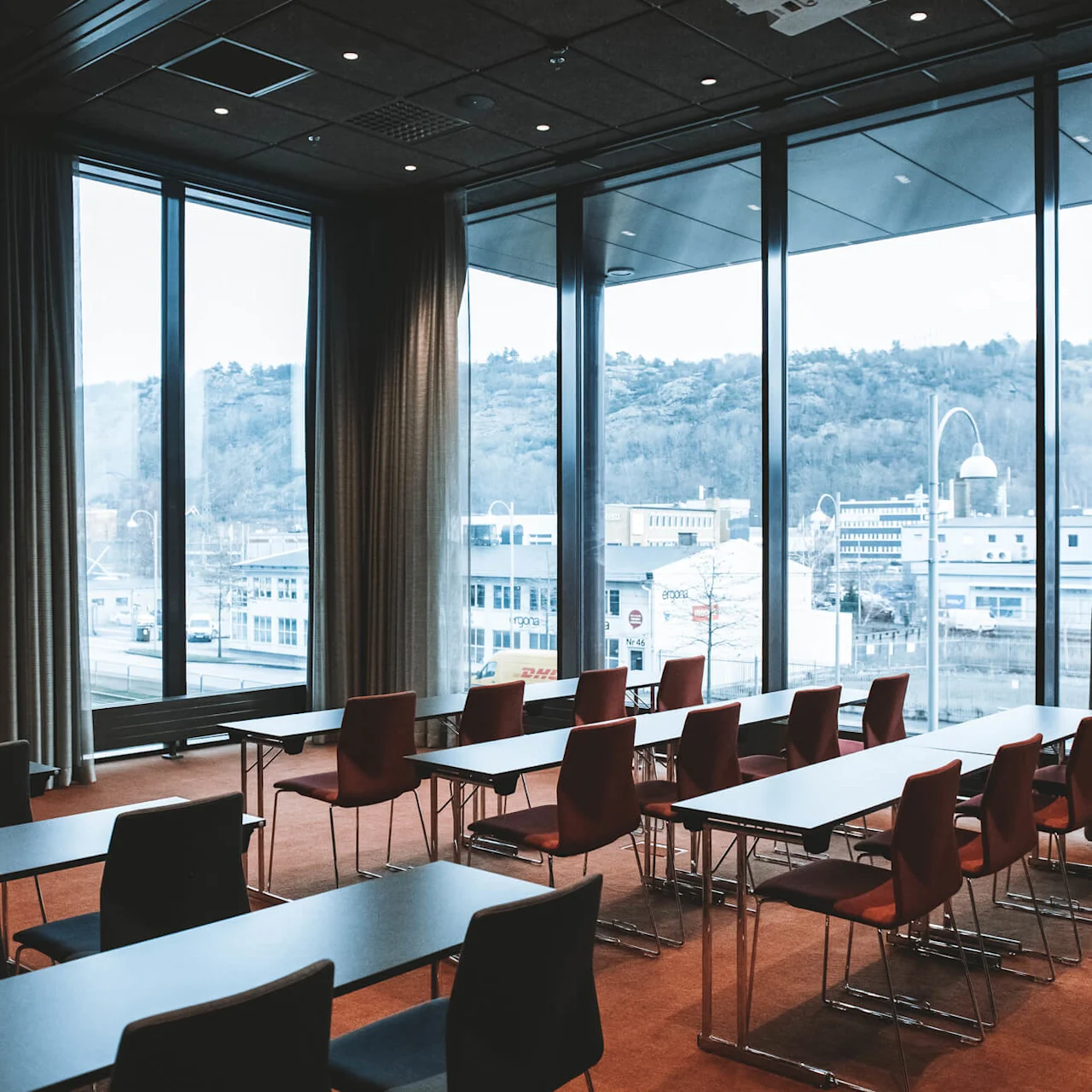 Konferensrum med tomma bord och stolar vända mot stora fönster. Utanför syns ett industriområde med byggnader och kullar. Rummet är väl upplyst, med gardiner delvis fördragna.