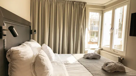 Ett ljust och inbjudande hotellrum med en bekväm säng med vita lakan, två badrockar och ett stort fönster.