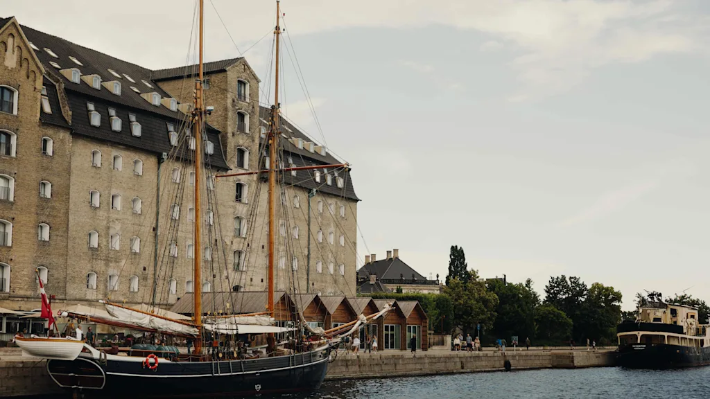 En historisk kanalscene med stor mursteinsbygning, seilbåt fortøyd og annen båt i det fjerne. Folk langs vannkanten.