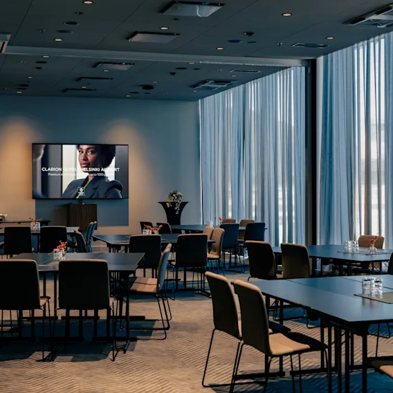 Kokoushuone Clarion Hotel Helsinki Airportissa, jossa pöydät ja tuolit on järjestetty konferenssia varten. Näytöllä lukee "HOTEL HELSINKI AIRPORT".