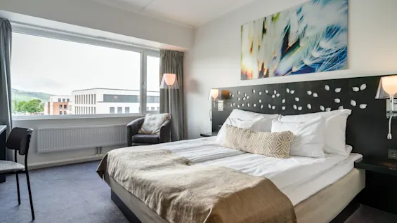 A neatly made bed with white linens in a modern hotel room. Large window with city view, two chairs, minimal decor, and soft lighting create a cozy atmosphere.