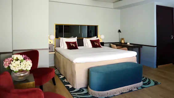 A neatly made bed with plush pillows stands in a modern bedroom featuring red chairs and a teal bench. A floral arrangement sits on a small table, enhancing the elegant decor.