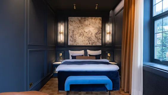 Elegant dark blue hotel room featuring a made double bed, stylish lighting, and a blue bench at the foot of the bed.