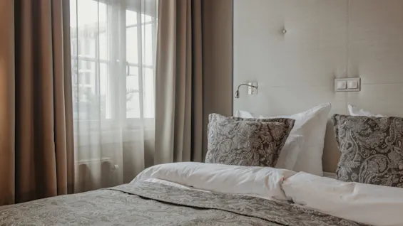 A serene hotel room with a comfortable bed, patterned pillows, a padded headboard, and a window with light filtering through curtains.