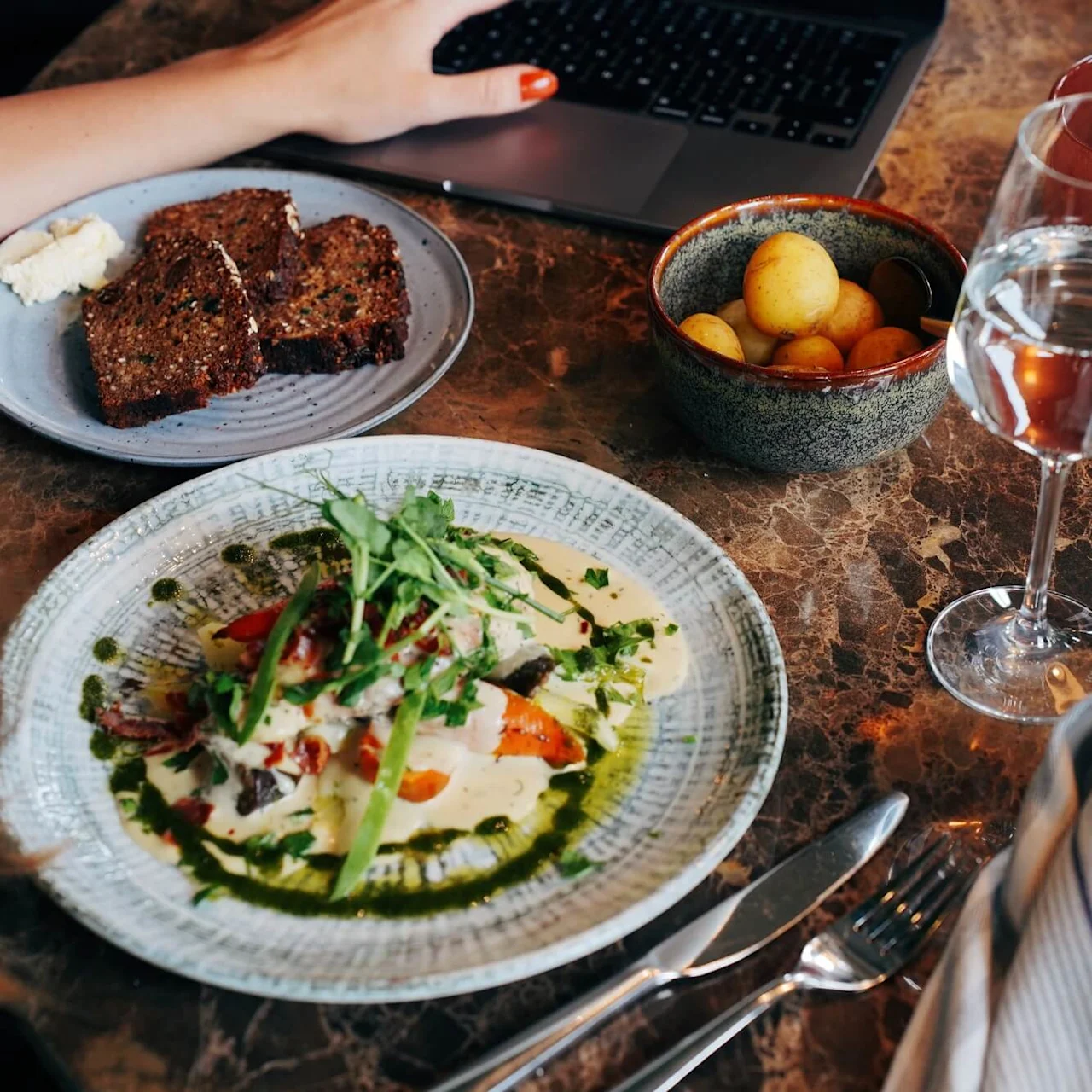 Person äter: en tallrik med sallad, bröd och en bärbar dator på ett marmorbord med ett glas vatten och vin.