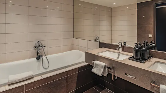 Bathtub with chrome fixtures and white towel, adjacent to a counter with dual sinks and black bottles, in a tiled bathroom.
