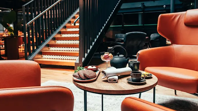 Et trebord holder tekanne, kopper, snacks i en koselig stue. Oransje skinnstoler omgir. Opplyst trapp i moderne, varmt opplyst interiør.