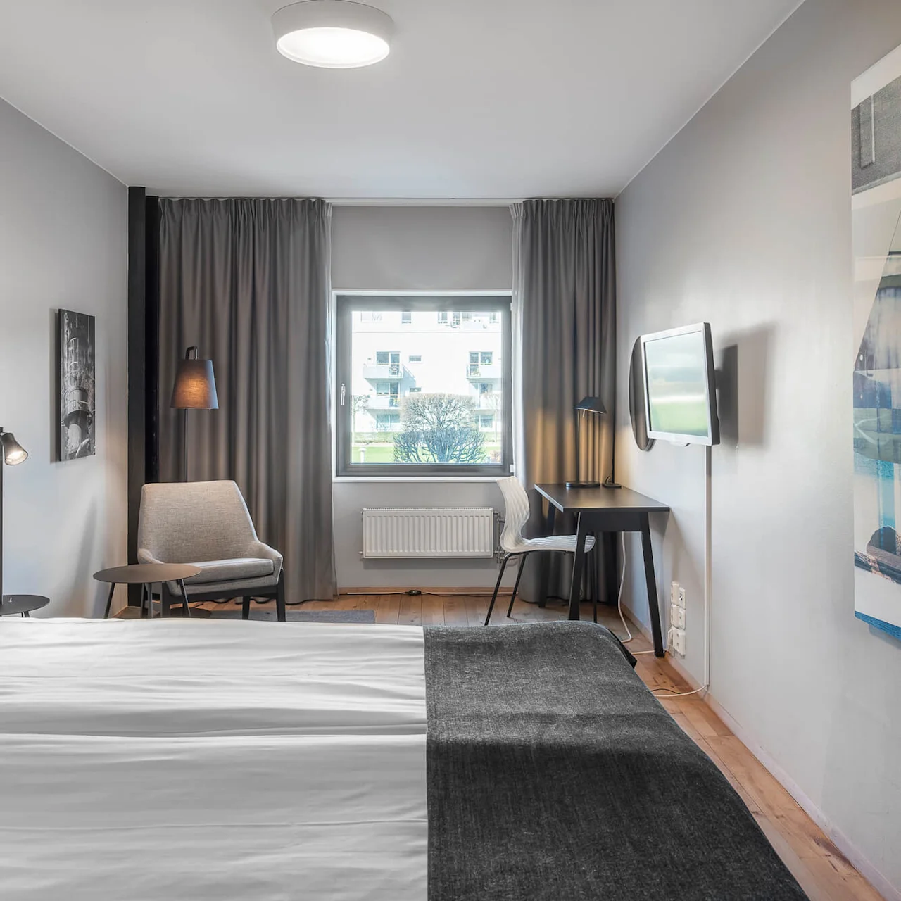 A modern hotel room features a neatly made bed, a small seating area with a chair and table, and a desk with a wall-mounted TV, all in a neutral-toned space.