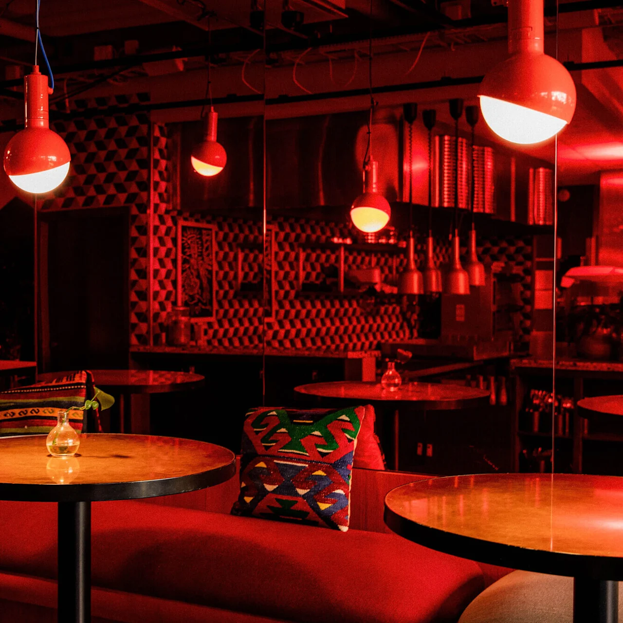 Red pendant lights illuminate a cozy, stylish lounge area with patterned cushions and wooden tables. Geometric wallpaper and modern decor create a warm, inviting atmosphere.