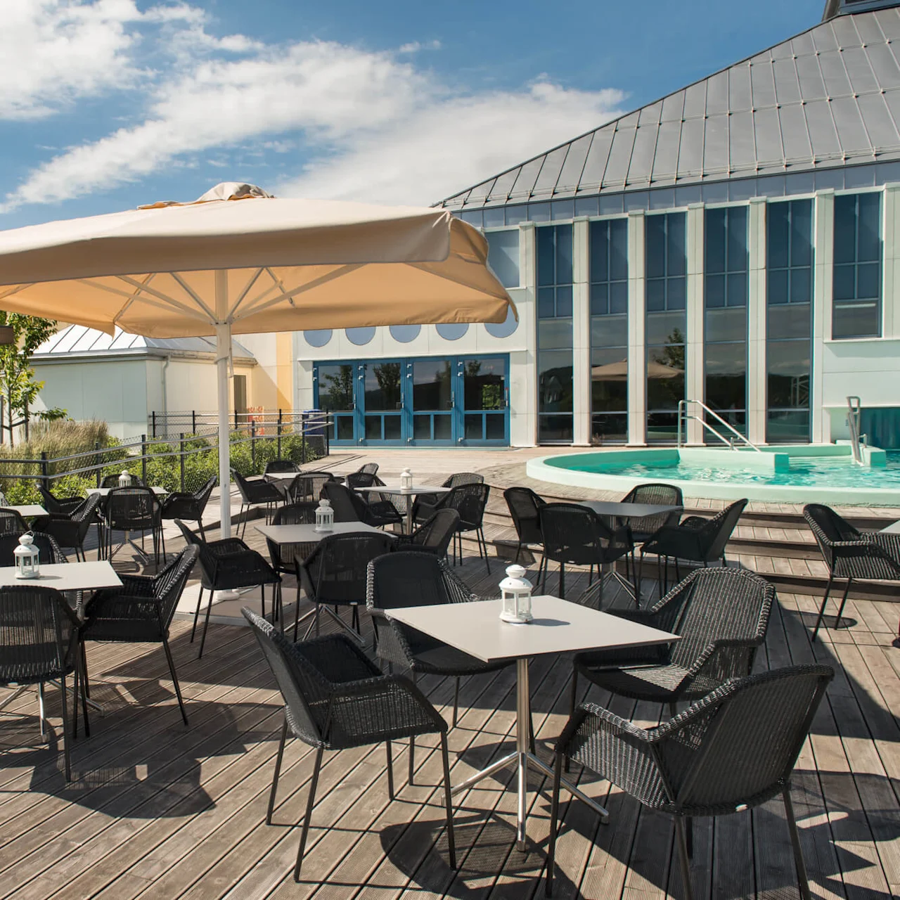Outdoor cafe tables and chairs sit under a large umbrella, near a wooden kiosk with drinks, adjacent to a modern building with glass windows and a small round pool.